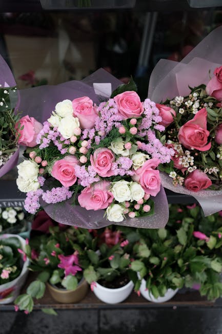 A floral bouquet featuring pink roses, white carnations, and small pink and white filler flowers, arranged with lush green foliage. The flowers are wrapped in soft purple tissue paper, with some in tr