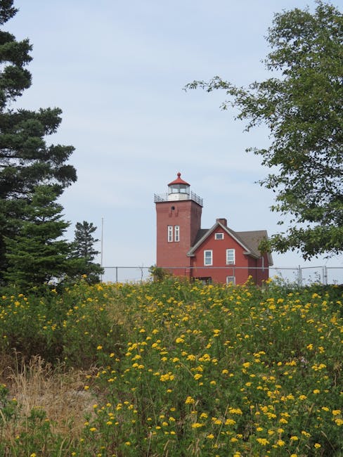 The image features a red brick lighthouse with a white lantern room and a small adjacent building, set against a partly cloudy sky. In the foreground, there is a vibrant field of yellow wildflowers an