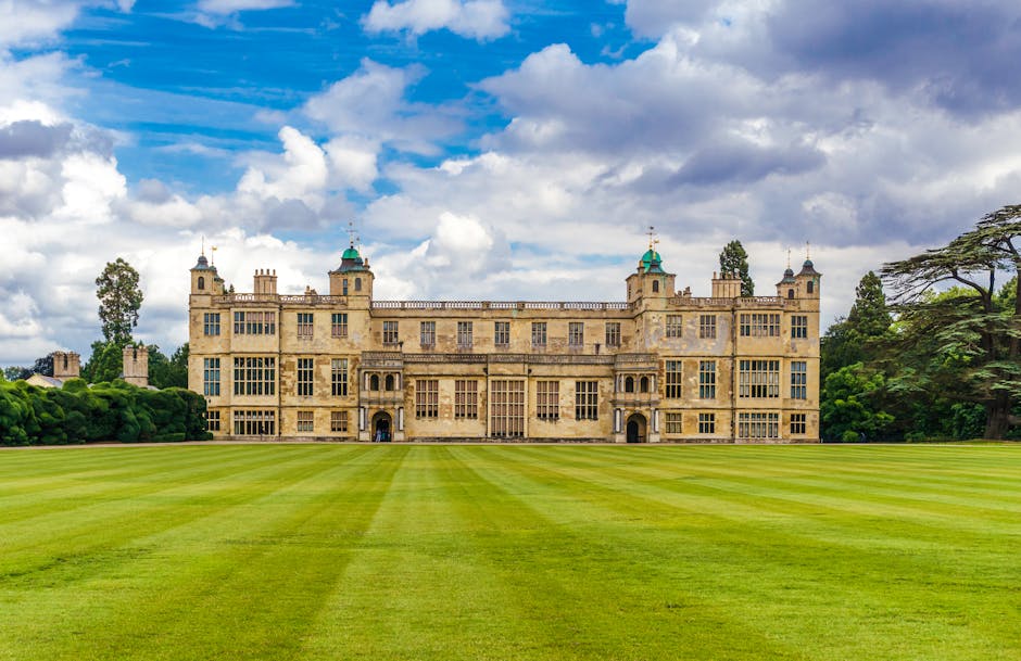 The image shows a grand historic castle with a symmetrical design, featuring a beige stone facade, multiple tall windows, and decorative towers with green domed rooftops. The castle is set against a d