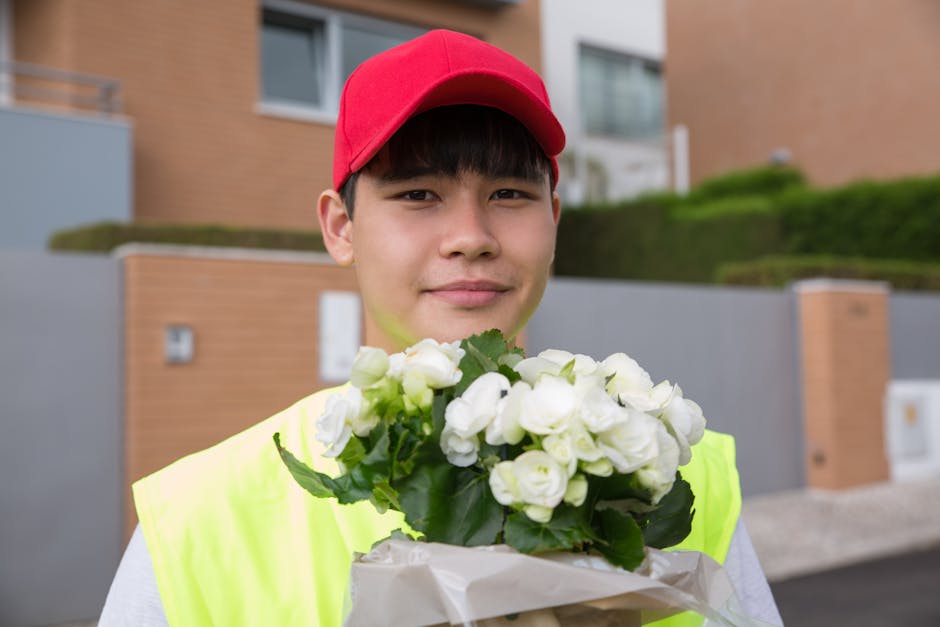 A young delivery person wearing a bright yellow high-visibility vest and a red cap is standing outdoors in front of modern residential buildings. They are holding a bouquet of fresh white roses with l