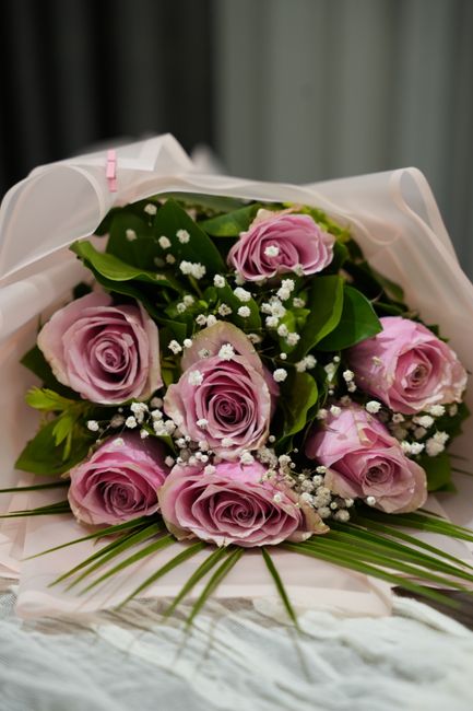 A bouquet of nine light pink roses with lush green leaves, accented by small white baby's breath flowers, arranged in a circular pattern. The flowers are wrapped in elegant white tissue paper with a s