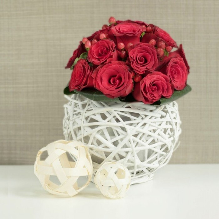 A carefully styled floral centerpiece, perfect for a wedding table in Charlton, dominates the image with its rich red hues and sculptural form. At the top of the arrangement, a compact dome of red roses is packed tightly together, their velvety petals layered in spirals that range from tightly wound buds to fully open, romantic blossoms. The reds are intense and saturated, with subtle variations of shades that create natural depth. Between the roses, fine stems of red hypericum berries peek through, their rounded, polished surfaces echoing the flower heads yet adding a slightly different texture, like tiny jewels set amongst silk.
The bouquet is supported by a hidden base of deep green foliage, which quietly frames the blooms and helps them stand out. These flowers are gathered into a striking spherical vase crafted from white-painted twigs or wicker, woven into an open, airy lattice. Through the gaps in the weave, glimpses of shadow and light play against the white, creating a modern rustic effect that would feel at home on a reception table in Charlton Village or in a smart dining room along Charlton Church Lane. The crisp white vessel accentuates the fiery reds of the roses, giving the arrangement a clean, contemporary profile.
Resting on a smooth white tabletop, the centerpiece is flanked on the left by two decorative wooden balls, fashioned from natural, light-toned strips delicately twisted and woven. The larger orb sits closer to the front, with the smaller one just beyond it, their understated beige colouring and organic texture gently balancing the vivid floral display. In the background, a softly blurred, neutral wall in beige or taupe-perhaps textured wallpaper or woven fabric-provides a calm, unobtrusive setting. The overall impression is one of elegance and intimacy, ideal for wedding receptions, anniversaries, or romantic dinners in Charlton.