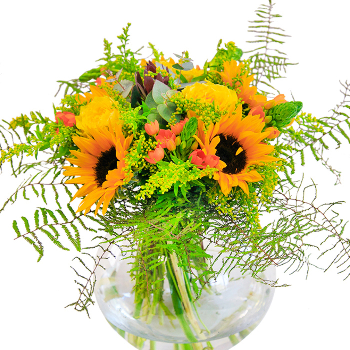From the heart of Charlton, this lavish bouquet is a vibrant ode to the sunniest days the area knows, arranged in a radiant, globe-shaped glass vase set against a clean white surface. Lush sunflowers form the soulful heart of the display, their wide, bright yellow petals circling velvety brown centers, just like blooms in a nearby allotment at peak season. Between them, opulent yellow garden roses spiral open, their intricate folds catching the light and drawing the eye-layers reminiscent of classic English gardens found off Charlton Road. Clusters of bold red hypericum berries are woven through the generous green foliage, their glossy hue providing lively contrast and whimsical charm. Fern fronds and lacy filler greens gracefully spill outwards, softening the silhouette and lending a sense of airy movement, as though ruffled by a gentle Greenwich breeze. Stems anchor elegantly in a crystal-clear, fishbowl-style vase, highlighted by the play of light on water and fresh, verdant lines. The fragrance, subtle yet uplifting, blends hints of cut wildflowers and sweet garden roses, evoking the promise of summer in every breath. Brimming with character and crafted with local flair, this arrangement is ideal for celebratory tables or thoughtful gifts, bringing a brilliant burst of Charlton's warmth and heart into any home or gathering.