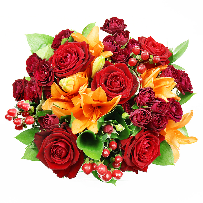 Directly photographed from above on a stark white surface, this opulent circular bouquet showcases a careful balance of colour, form and texture-an arrangement I often bring together when filling orders for Charlton neighbours around the train station. Several large, richly hued red roses take center stage with their velvety, multi-layered petals unfurling toward the viewer, evoking passion and a timeless elegance. Interlaced are bright orange Asiatic lilies whose trumpet profiles and visible stamens create striking sculptural accents; the lilies vary from fully open, dramatic blooms to elegant buds just beginning to reveal themselves, lending a sense of progression and life. Tucked into the spaces between are clusters of deep burgundy spray roses, their miniature, densely packed blooms adding luxuriant texture and deep tonal contrast. Scattered throughout, glossy Hypericum berries in a vibrant red introduce shimmering points of interest that catch soft studio light. Generous green foliage-broad, glossy leaves suggestive of ruscus or salal-peeks through and frames each blossom, giving the composition a fresh, garden-picked quality. The overall effect is one of sumptuous celebration: visually rich, layered and inviting, with the lilies' spicy citrus top notes and the roses' warm, honeyed depth almost perceptible. This bouquet sits perfectly on a white mantel or dining table and communicates affection, congratulations or thanks-an ideal floral choice for anniversaries, heartfelt apologies, or a welcome-home gift after a weekend in Greenwich Park or an afternoon along Woolwich Road.