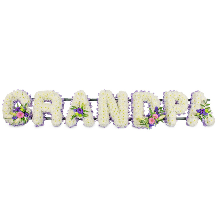 Set against a bright, uncluttered white backdrop, this floral tribute spells out GRANDPA in sturdy, upright capital letters, designed as a tender farewell for a beloved grandfather. Each letter is richly covered in small, snowy-white flowers, most likely chrysanthemums or spray carnations, placed so closely together that they form a plush, cushion-like surface. This dense white base gives the arrangement a look of purity and peace, echoing the quiet reflection often felt at funerals and remembrance services around Charlton. Tracing the outline of each letter is a fine border of dainty lilac and purple blooms, perhaps asters or statice, which gently frame the shapes and add subtle definition while maintaining a soothing, understated palette. Interspersed across the tribute are several thoughtfully composed accent sprays: the 'G' holds a cluster of bright pink blossoms, a refined white lily-like flower, and fresh green foliage; the 'D' mirrors this with a similarly graceful grouping of pinks, white, and greenery. The first and last 'A' each bear smaller tufts of rich purple florets with glossy dark green leaves, adding depth and visual rhythm along the word. A slim, dark green frame bar runs horizontally behind the letters, mostly hidden by petals, indicating that the piece is mounted for secure display, perhaps at a graveside gathering after a walk from Charlton station or a local funeral home. Soft, even lighting in the photograph gently illuminates every petal and curve, emphasising the careful craftsmanship and the emotion invested in this personalised memorial flower arrangement for Grandpa.
