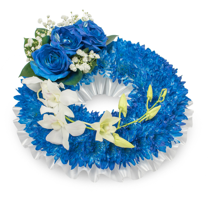 A dignified, circular wreath presented on a neutral white backdrop, carefully assembled to balance deep, contemplative blues with strokes of pure white. The wreath's broad foundation is made up of innumerable royal-blue chrysanthemum-like blooms-each small, densely packed flower contributing to a richly textured ring that reads as both plush and solemn in the diffused studio light. A soft, ruffled white satin ribbon lines the inner circumference, its pleats and subtle lustre creating a refined border that frames the blue mass and suggests ceremonial formality. At the upper-left curve, a cluster of three magnificent blue roses commands attention; their petals are velvety and sculptural, unfurling in a deep sapphire hue that harmonises with the chrysanthemums while offering a distinct focal point. Around this rose cluster, fine sprays of white baby's-breath add a filigree of lightness and the occasional dark green leaf injects natural contrast. From the mid-left the arrangement is adorned by a cascade of pristine white orchids-likely Phalaenopsis or similar-whose broad, silky petals arc gracefully toward the lower right, creating a soft, flowing line across the wreath. Interspersed with the open orchids are fresh green buds, some taut with potential, suggesting renewal threaded through the act of remembrance. The overall impression is one of solemn beauty and quiet comfort: the blue elements evoke memory and reverence, the white orchids and ribbon name purity and peace. Locally, this style of wreath is often chosen for memorial services held near Charlton Park, riverside commemorations, or as a respectful display delivered to chapels and funeral directors serving Charlton and neighbouring Greenwich.