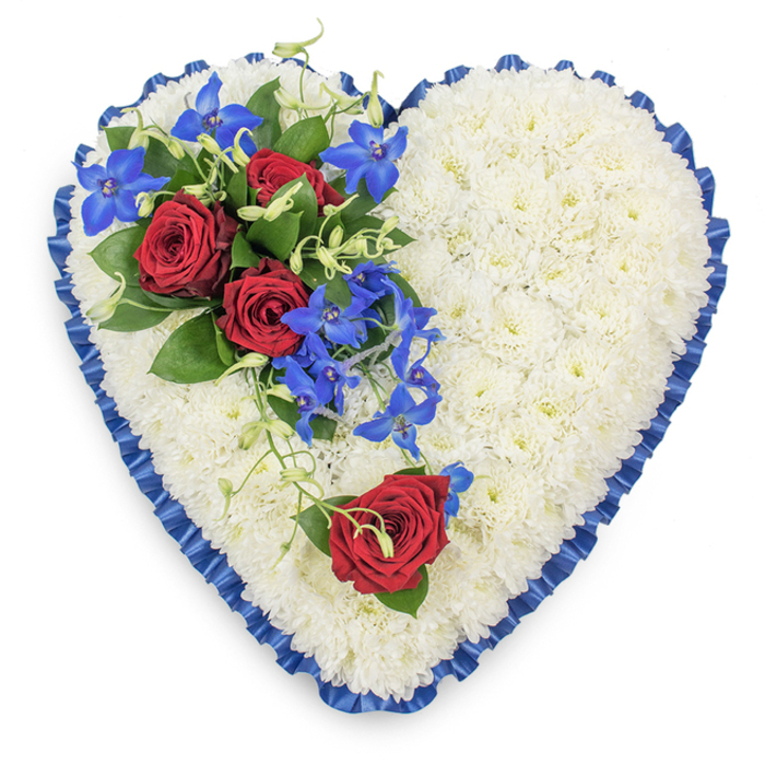 In this image, a generously sized heart-shaped funeral tribute is shown from a top-down perspective, allowing every nuance of the floristry to be appreciated. The body of the heart is formed entirely from compact white chrysanthemums, the small spray blooms arranged so closely that they create a thick, plush surface, soft and immaculate, evoking purity and quiet remembrance. Along the outer edge, a royal blue satin ribbon has been meticulously gathered and pleated, its rippled texture and glossy finish creating a decorative frame that both outlines and elevates the shape. Positioned on the upper left of the heart, a carefully composed cluster of flowers introduces a vivid burst of colour: three deep red roses, fully opened with velvety petals curling outward, rest among rich, dark green leaves that add depth and structure. Threading through this cluster, bright blue dendrobium orchids arc downwards, their distinctive star-shaped petals shimmering in intense cobalt and teal tones, while light green curly tendrils weave in and out, softening the lines and lending a natural, flowing feel. On the lower right curve, a solitary red rose with a small accent of foliage sits slightly apart, echoing the main spray and adding emotional resonance, like a final whispered farewell. The tribute feels calm yet profoundly expressive, ideal for a funeral service or wake in Charlton, whether placed beside a coffin in a local church or carried from a home near Charlton Park to the crematorium. Designed with care typical of Flower Delivery Charlton, it captures the balance of devotion, respect, and serenity that families often seek at moments of loss.