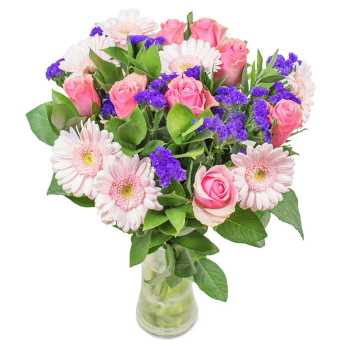 An artful floral arrangement stands as a centrepiece in a clear glass vase, illuminated against a pristine white backdrop that could be found in a welcoming Charlton home. Approximately seven to nine medium pink roses, each with lush, spiralling petals at stages from slightly furled to just beginning to open, anchor the bouquet with timeless beauty and romance. Interspersed among them are five to seven pale pink gerbera daisies, recognisable by their broad, delicate petal displays and pale, nearly white centres, radiating a gentle warmth. Clusters of vivid, deep purple statice peek through the pink blooms, adding striking contrast and a tactile, textured flourish that enlivens the palette. Rich, emerald green foliage cradles the blossoms, overflowing with freshness and vibrancy, suggestive of a walk through Charlton's inviting green spaces. The arrangement is completed by a slender, transparent vase showcasing sturdy stems and clear water, reinforcing the sense of vitality and immediacy typical of a freshly delivered bouquet from a reputable local florist. This bouquet-ideal for birthdays, anniversaries, and heartfelt celebrations-conveys a message of joy, tenderness, and the particular charm of gifting flowers in Charlton, offering a delightful visual and aromatic presence that brightens both hearts and rooms.