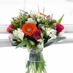 In this captivating Charlton-inspired floral arrangement, artistry and nature unite in a lush, radiant bouquet that feels both sophisticated and inviting. At its heart, creamy gerbera daisies form a delicate circle, their soft petals radiating from deep brown centres that glow with the warmth of autumn sunsets seen from Charlton Park. Brilliant orange gerberas add flashes of zest, while dramatic magenta roses inject a note of romance and luxury amid the ensemble. Pure white alstroemeria, perhaps mingling with dainty spray roses, lend a fresh counterpoint-a whisper of possibility and new beginnings. Tiny, pale pink accents and minute filler blooms nestle between the larger flowers, providing layers of texture and nuance. Enclosing the stems, large aspidistra leaves arc gracefully, forming a polished, structured base that underscores the arrangement's full, rounded shape. The display feels rooted in the landscape, as smaller, pointed green leaves thread through, reminiscent of the greenery found along the Thames Path nearby. All stems are showcased in a tall, clear glass vase, letting the vibrant greens gleam in the light and emphasizing the freshness so valued by the Charlton community. The soft, white background invites attention to the joyous palette and lush textures, while a gentle, garden-like fragrance lingers in the imagination. This bouquet is a reflection of celebration, warmth, and local pride, ideal for birthdays, housewarmings, or meaningful gatherings in SE7-crafted with expertise worthy of Flower Delivery Charlton.
